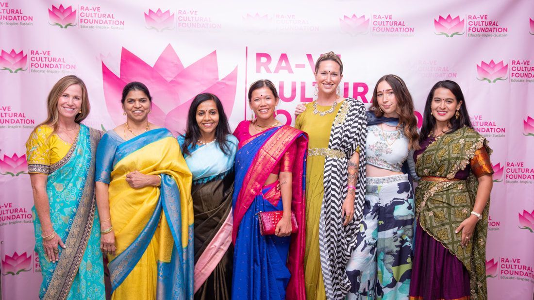 Group of women posing, wearing sarees and dresses, in front of a pink backdrop with lotus flower logo.