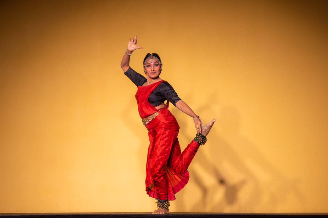 A dancer in red costume performs against a yellow backdrop, one arm raised, foot lifted.
