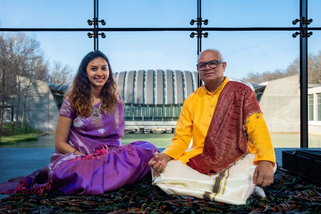 Woman and man sit cross-legged, holding hands. Outdoors with modern building backdrop.