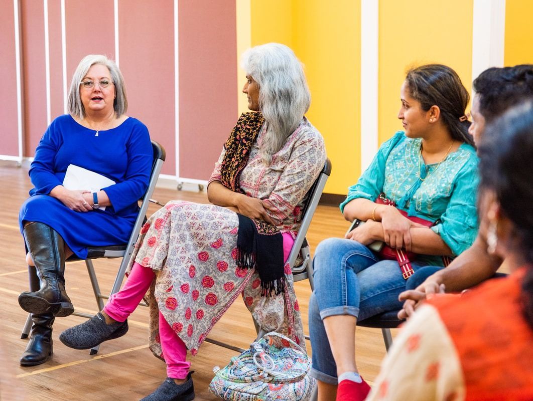 People seated in a circle, engaged in a discussion. Woman in blue, one in floral print. Well-lit room.