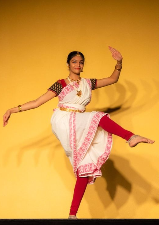 Woman in white and red Indian dance attire, poised mid-step, right leg raised, arms curved, against yellow background.