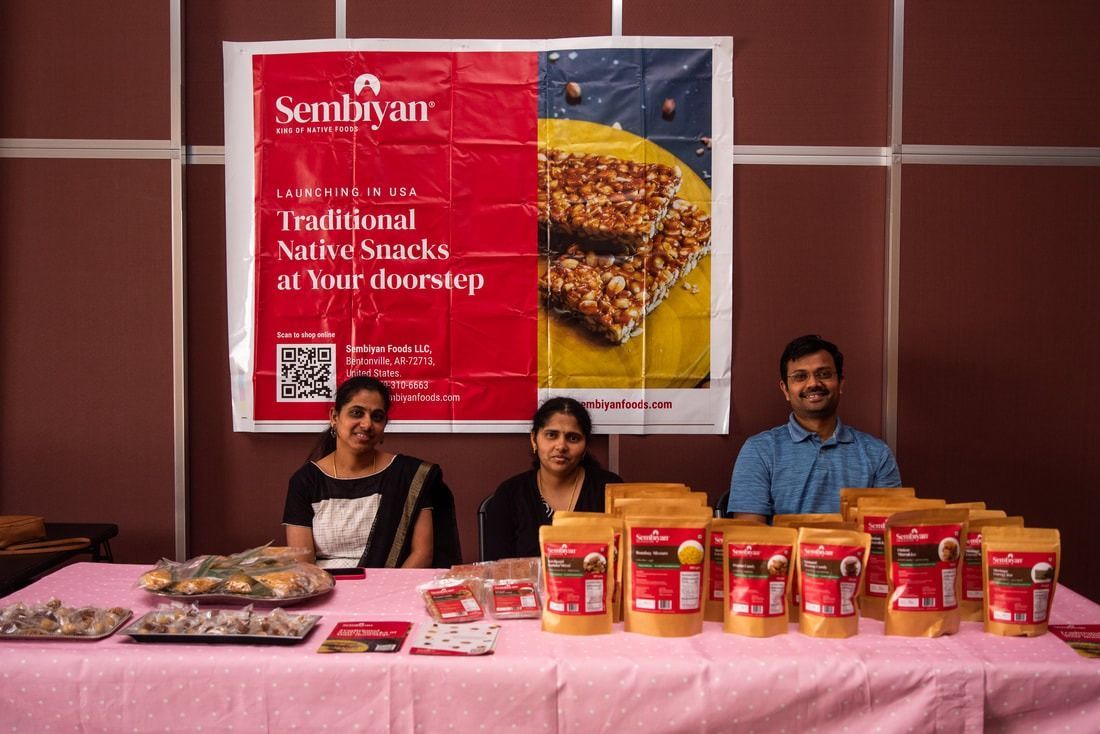 People at a snack booth with a red sign and packages of snacks on a pink table.