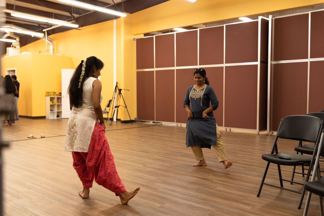 Two dancers in a studio with a tripod and chairs, one in white and pink, the other in blue, performing.