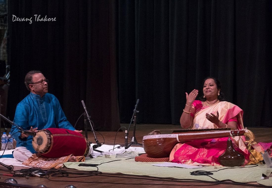 Musicians perform on stage; a woman sings and gestures, a man plays a drum-like instrument.