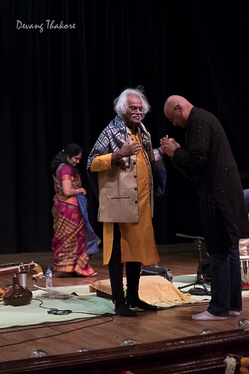 Man in yellow outfit and another man bowing on stage; woman in sari in the background.