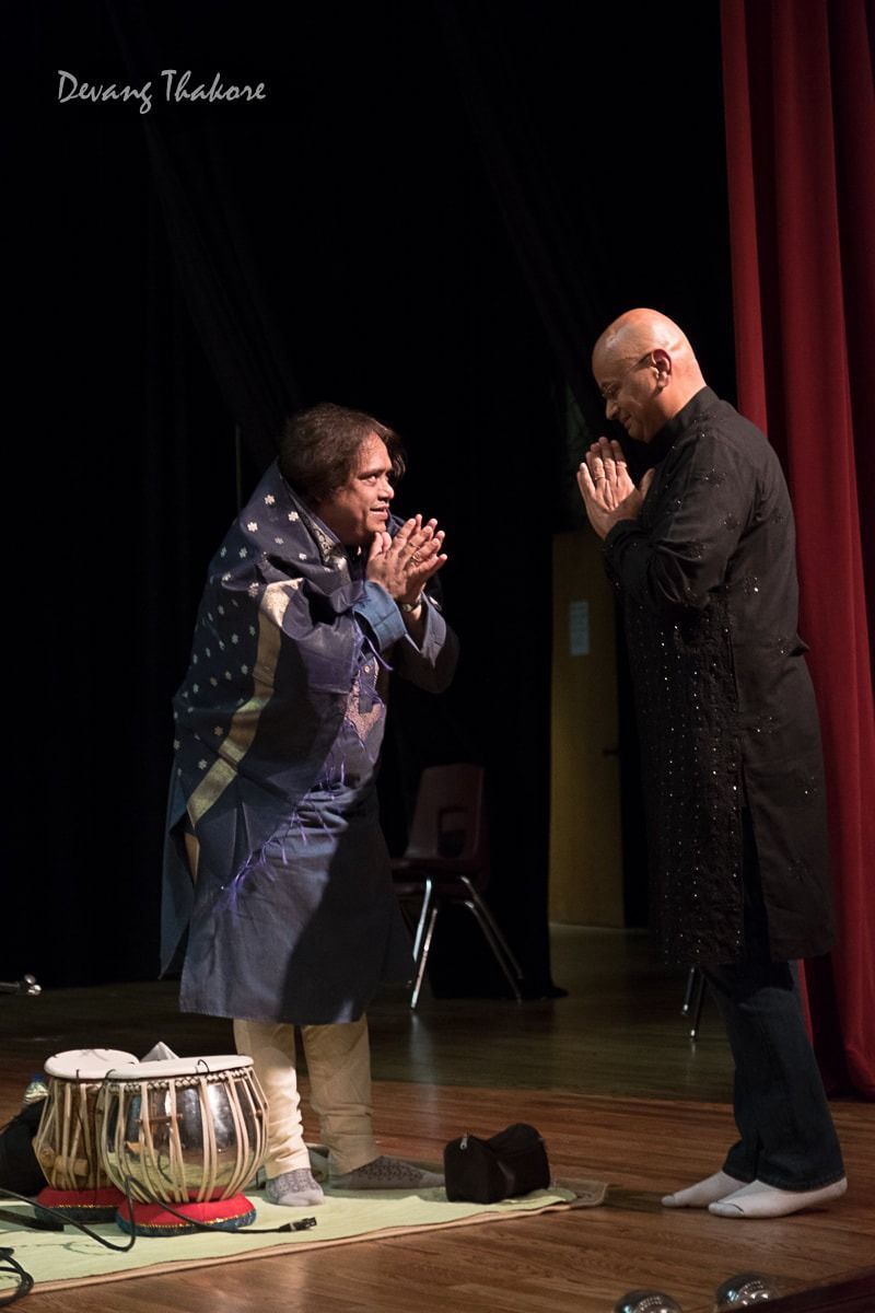 Two people on stage with hands pressed together, facing each other. A tabla drum sits nearby.