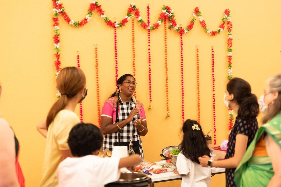Woman in apron speaks to a group, garland backdrop, food on table.