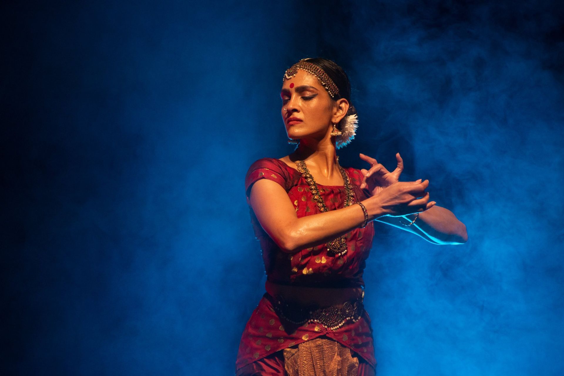 Woman in red dance attire, performing Indian classical dance; blue, smoky background.