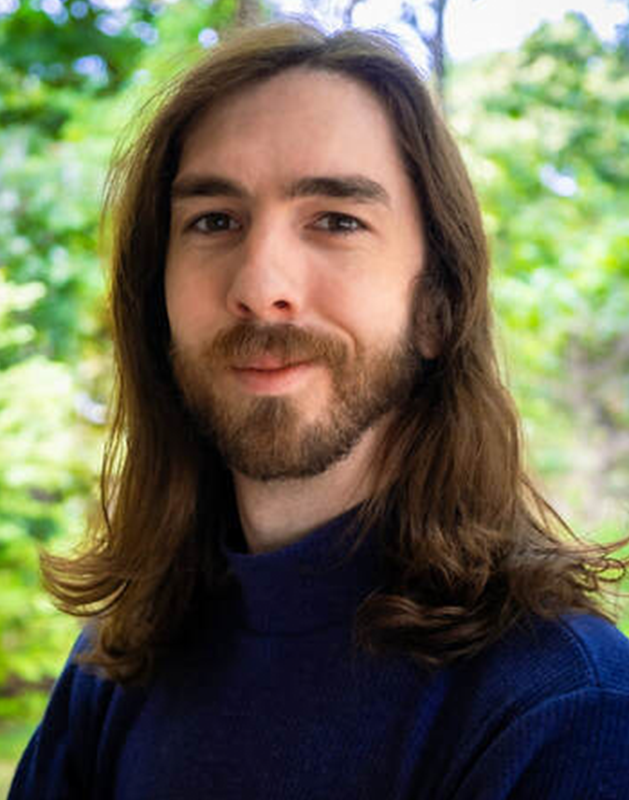 Man with long brown hair and beard smiles, wearing a blue turtleneck, outdoors in a green, leafy environment.