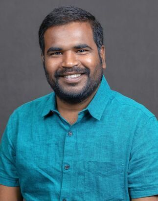 Man with dark hair and a beard, smiling, wearing a blue button-down shirt.