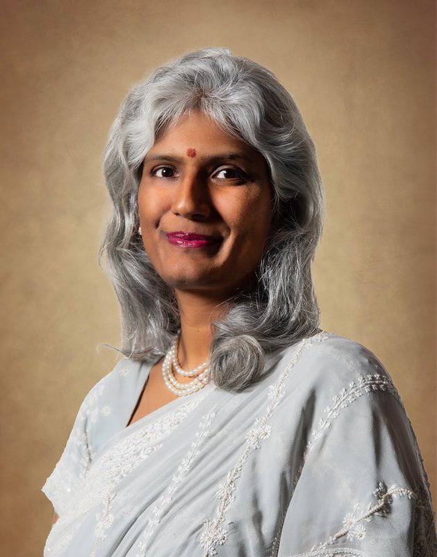 Woman with gray hair, wearing a light blue sari and pearl necklace, smiles.
