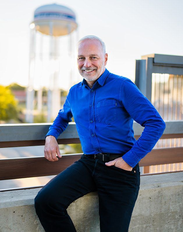 Man in blue shirt and black pants leans on a railing, smiling with a water tower in the background.