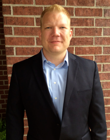 Man in blue shirt and navy blazer smiles in front of a brick wall.