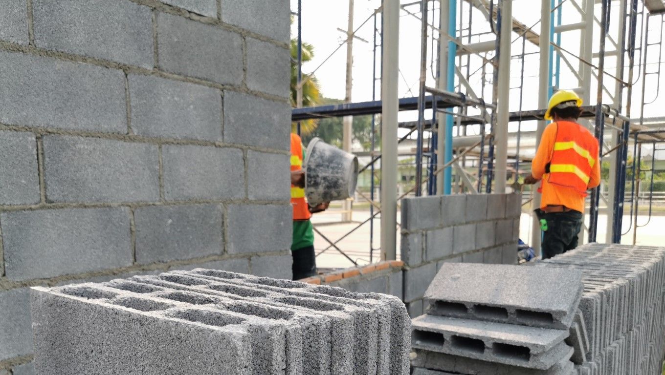 Construction workers in orange vests building a cinder block wall at a site.