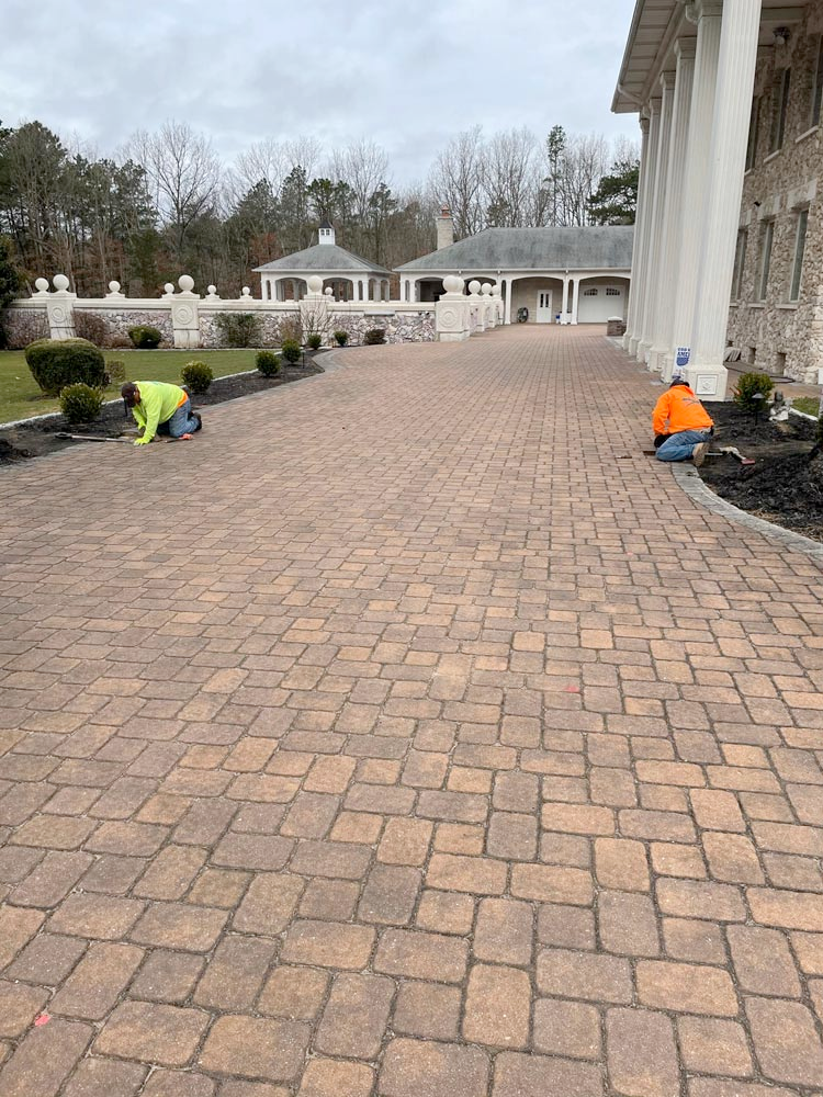 Two men are working on a brick driveway in front of a large house.