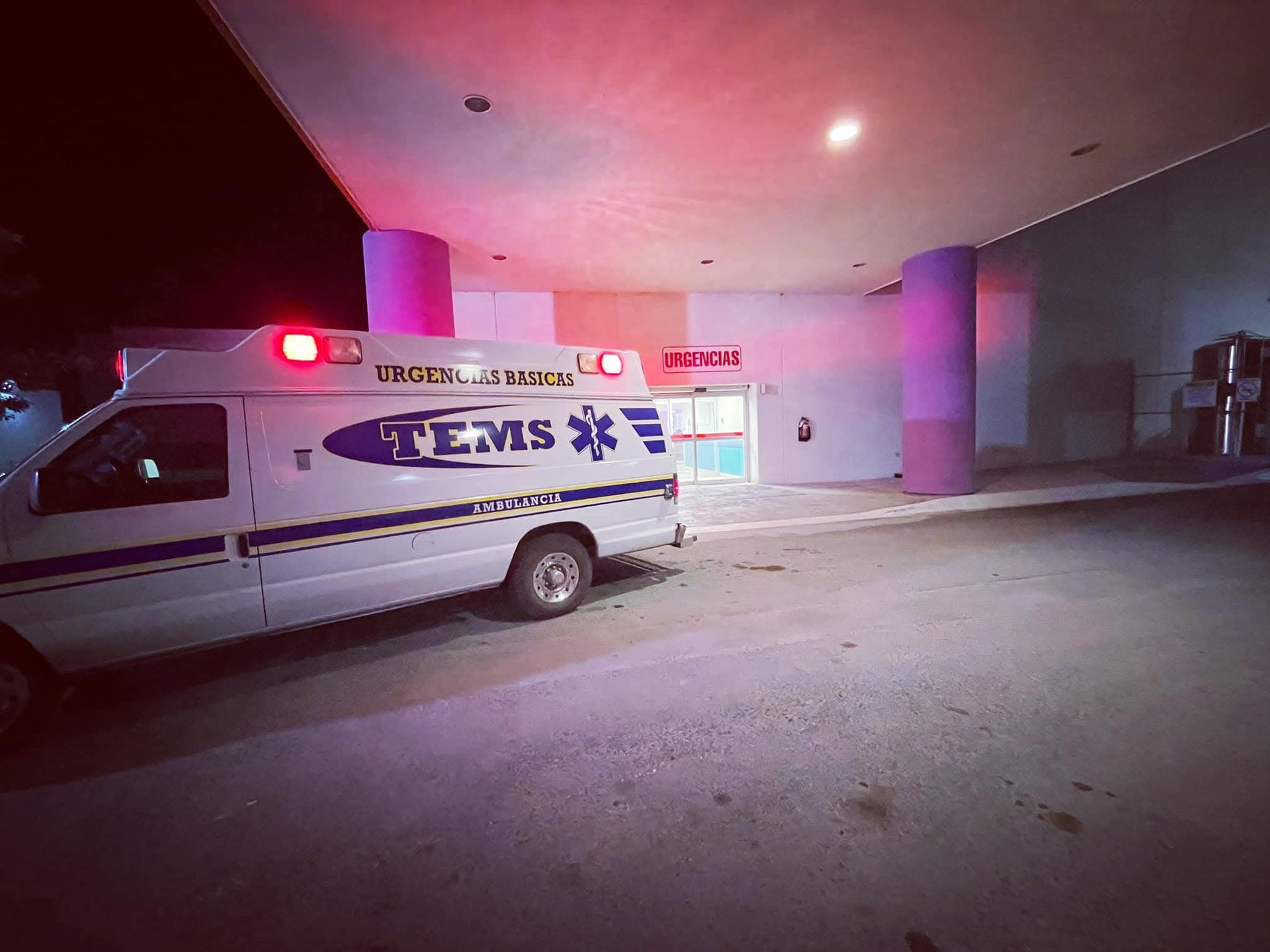 Ambulancia estacionada en la entrada de un hospital con luces rojas intermitentes; escena nocturna.