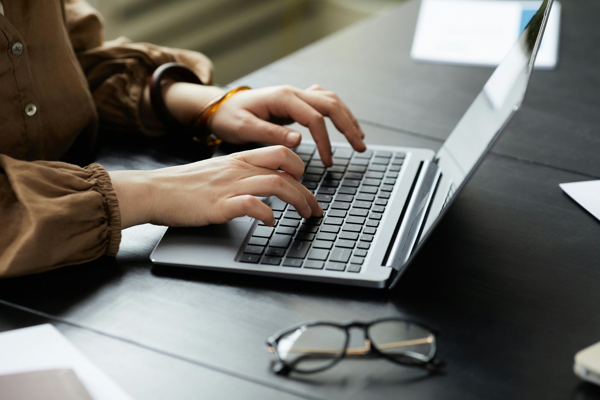 Person typing on laptop at a desk. Hands are visible; glasses and papers on the desk.
