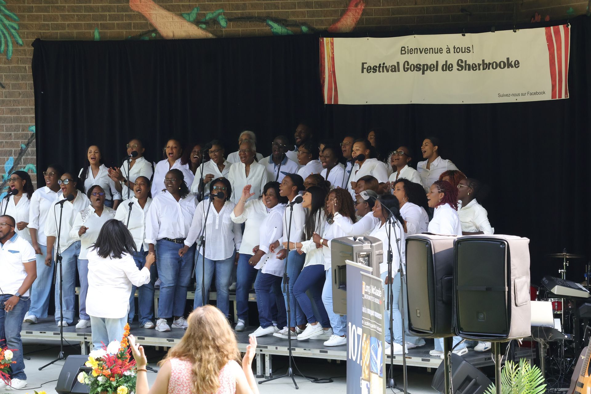 Chorale sur scène, chantant en plein air, vêtue de chemises blanches et de jeans. Une femme dirige.