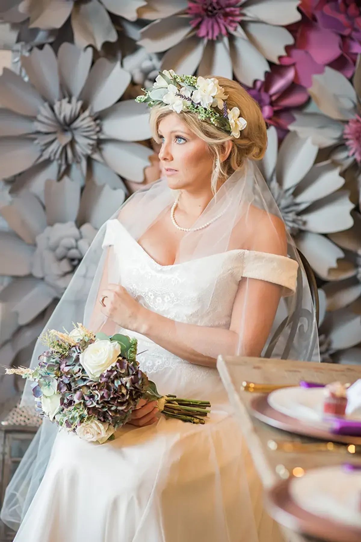 A bride is sitting in front of a wall of paper flowers holding a bouquet of flowers.