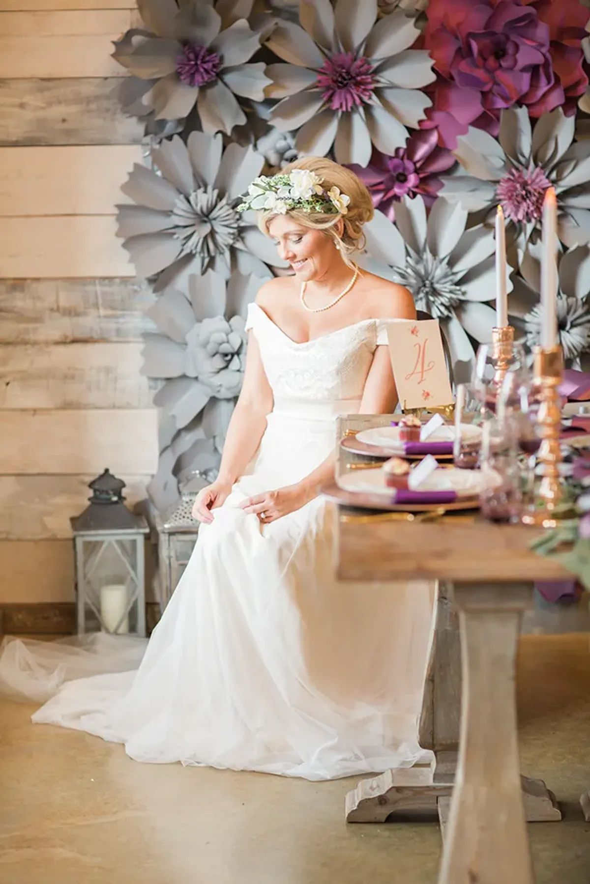A bride in a wedding dress is sitting at a table in front of a wall of paper flowers.