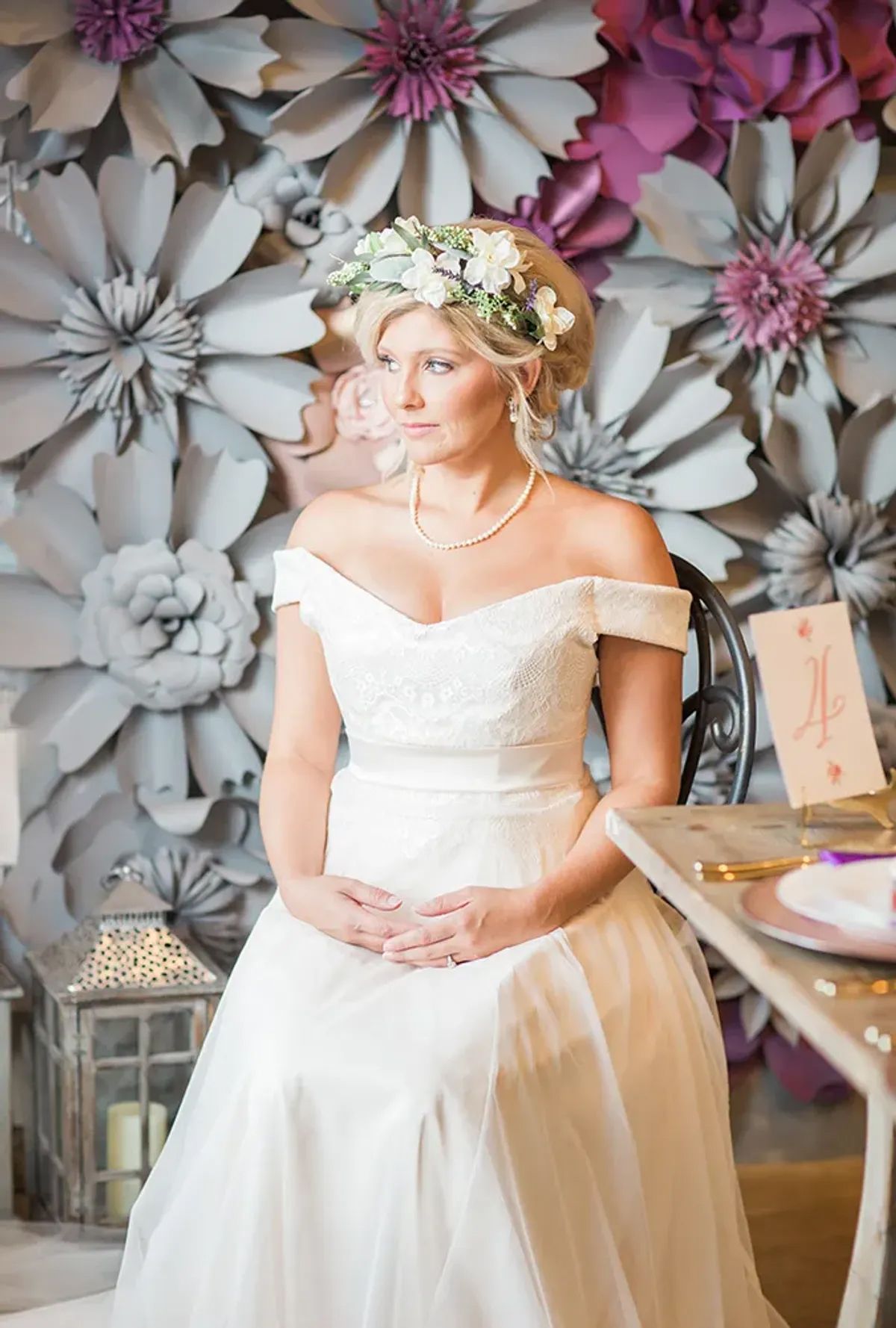 A woman in a wedding dress is sitting at a table in front of a wall of paper flowers.
