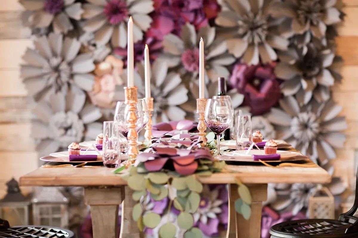 A table with plates , candles , and flowers on it in front of a wall of paper flowers.