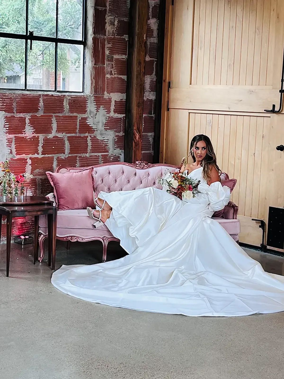 A bride in a long white dress is sitting on a pink couch holding a bouquet of flowers.