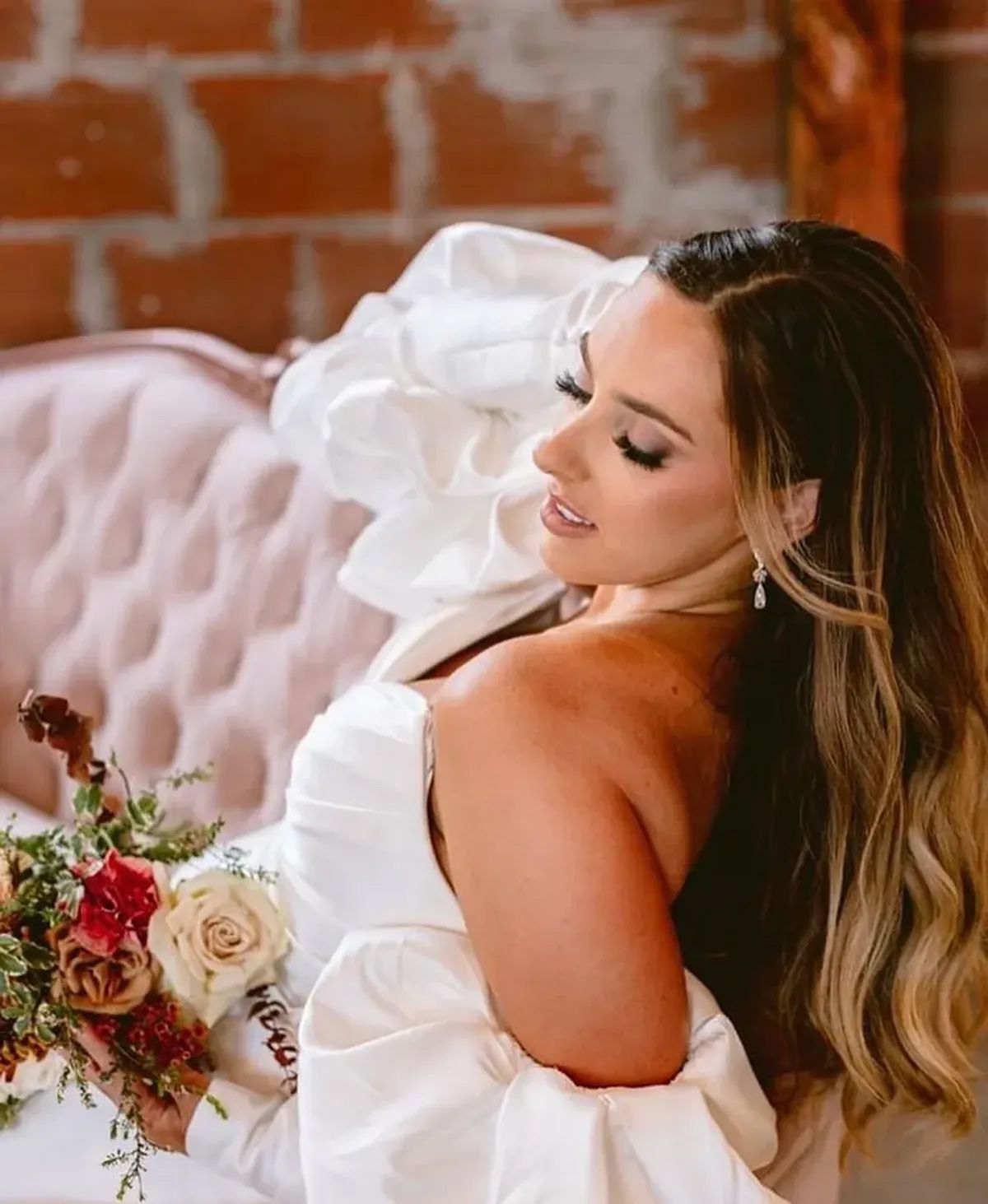 A woman in a white dress is sitting on a pink couch holding a bouquet of flowers.