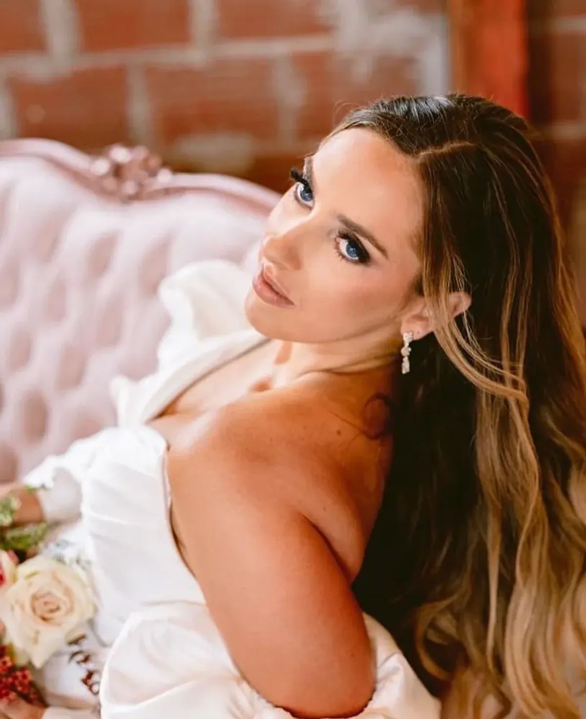A woman in a white dress is sitting on a pink couch holding a bouquet of flowers.