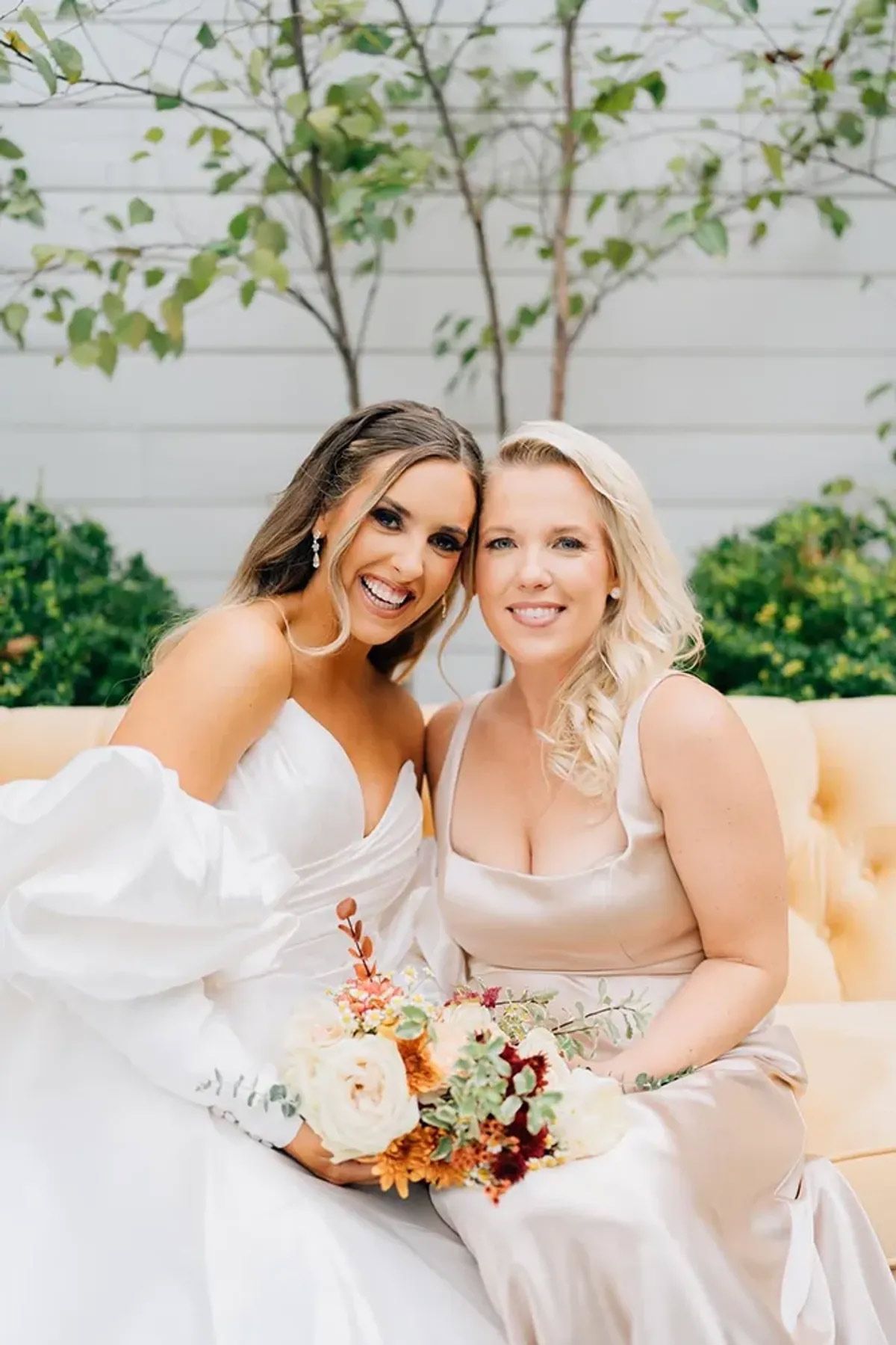 A bride and her bridesmaid are posing for a picture while sitting on a couch.
