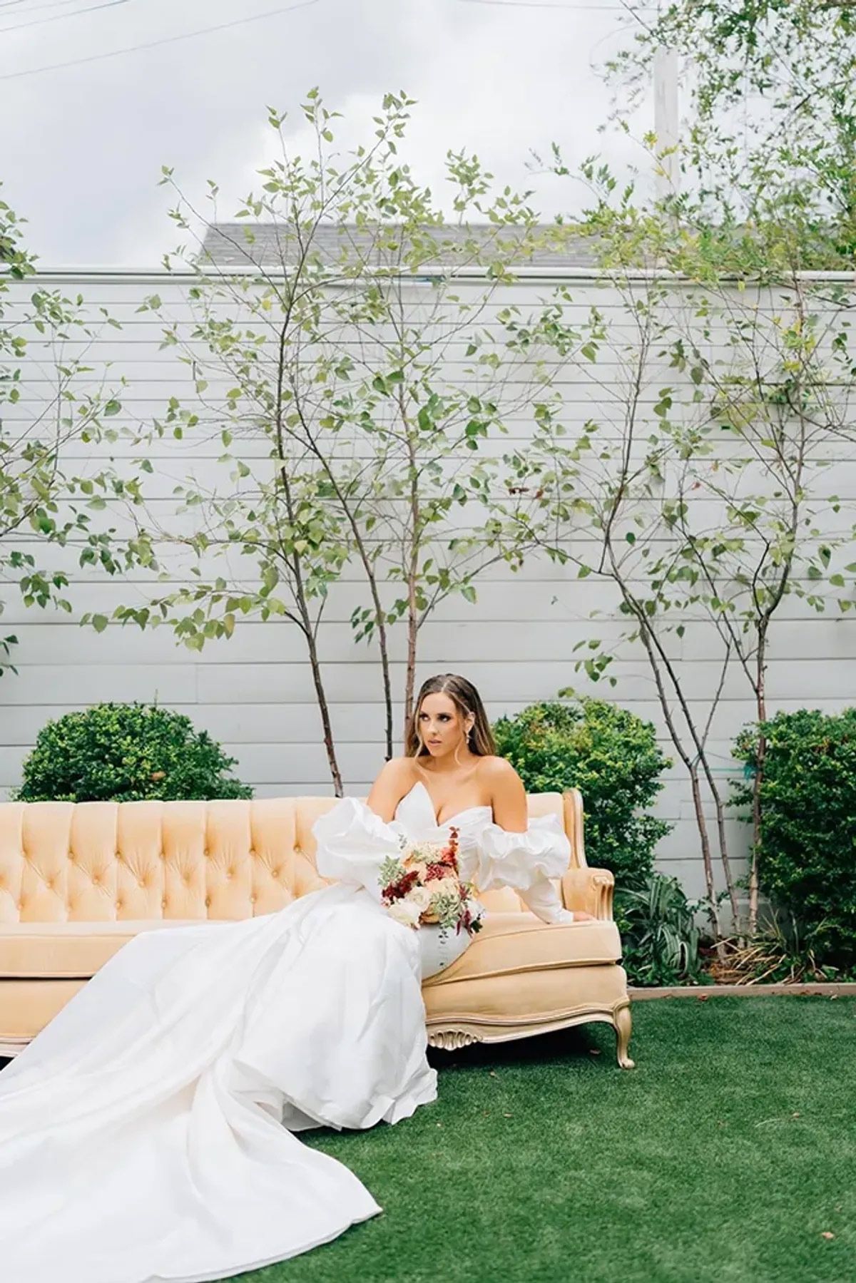 A bride in a white dress is sitting on a couch in the grass.