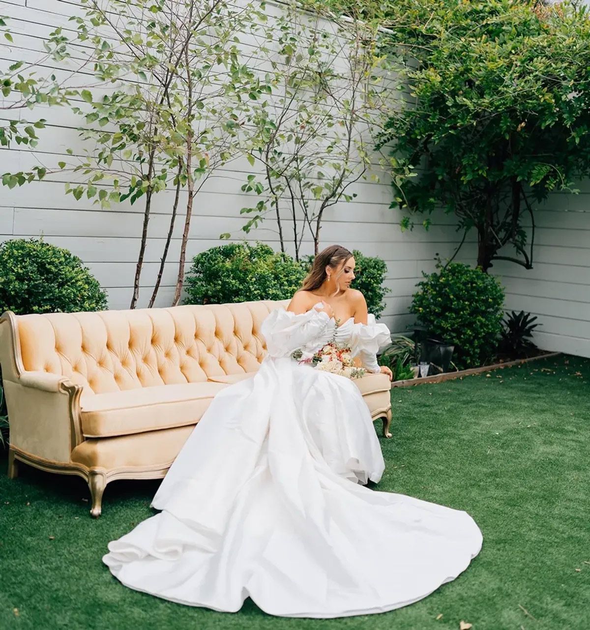 A woman in a wedding dress is sitting on a couch in the grass.