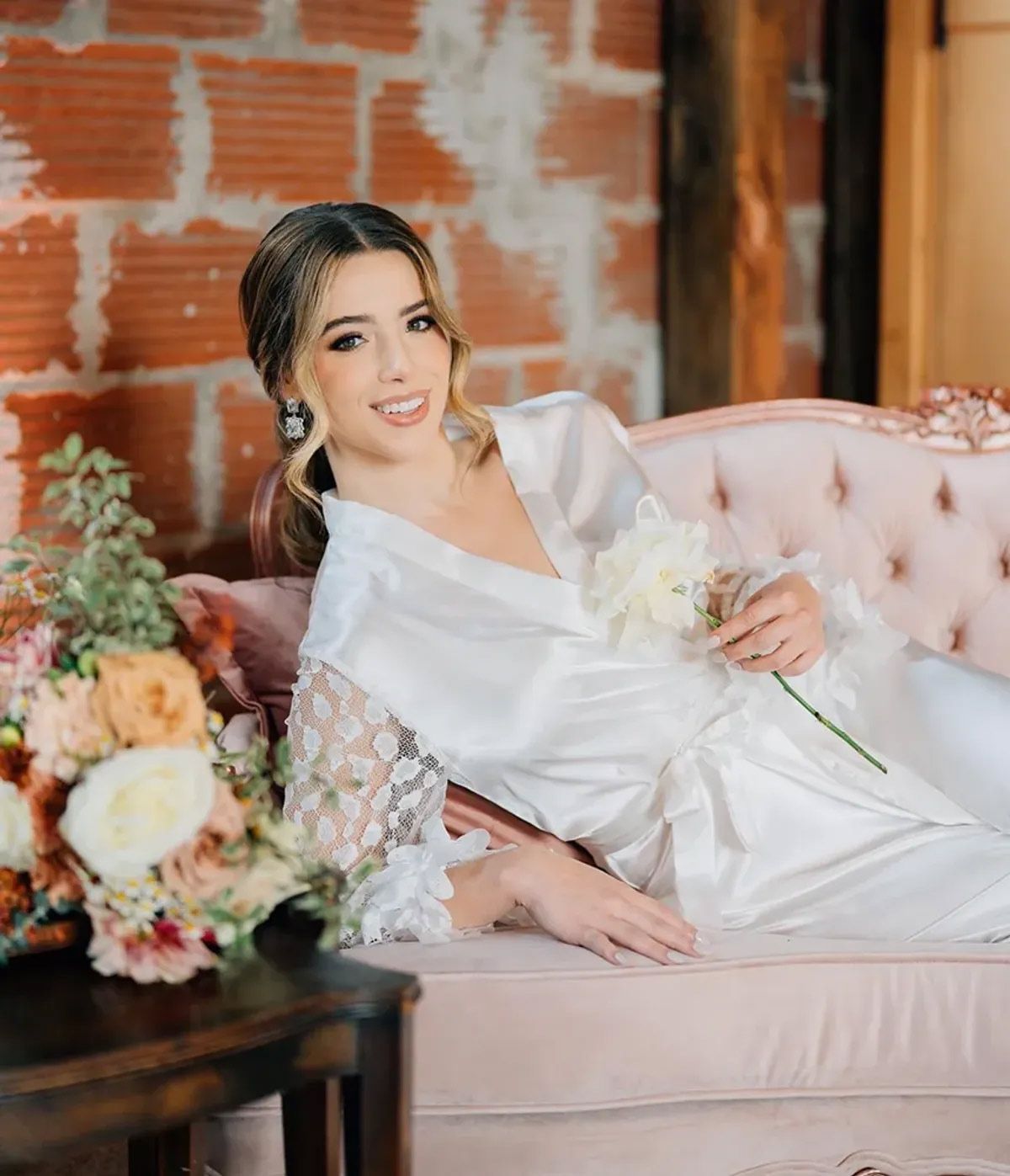A woman in a white robe is sitting on a pink couch holding a bouquet of flowers.