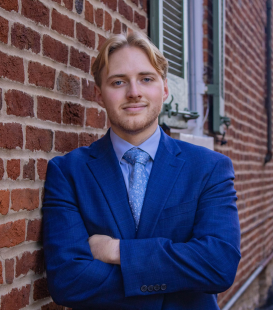 Blond man in blue suit leans against a brick wall with arms crossed, smiling.