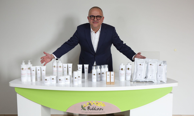 Una persona in giacca e cravatta si trova dietro un tavolo bianco e verde che espone una collezione di flaconi e astucci bianchi per prodotti per la cura della pelle.