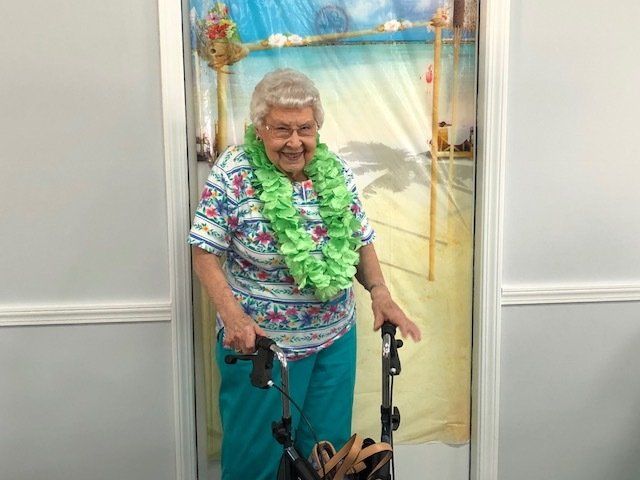 An elderly woman wearing a lei and a walker is standing in front of a door.