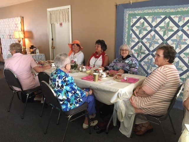 A group of people are sitting around a table with a quilt in the background
