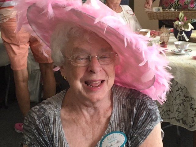 An elderly woman wearing a pink hat and glasses is smiling.