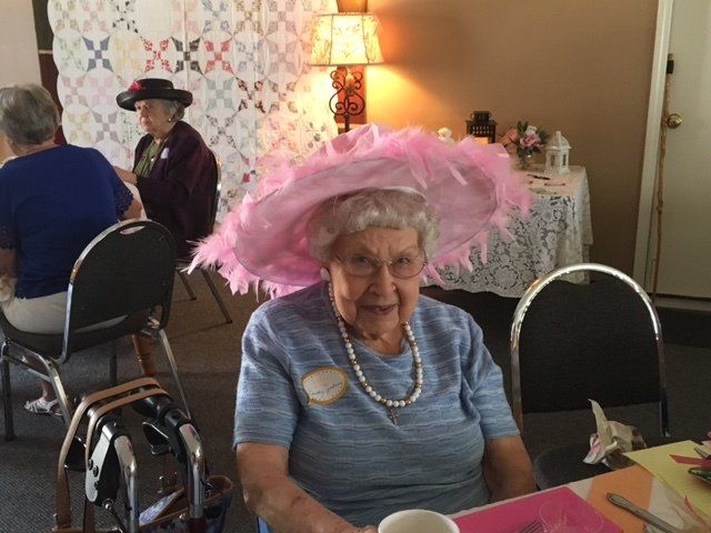 An elderly woman wearing a pink hat sits at a table