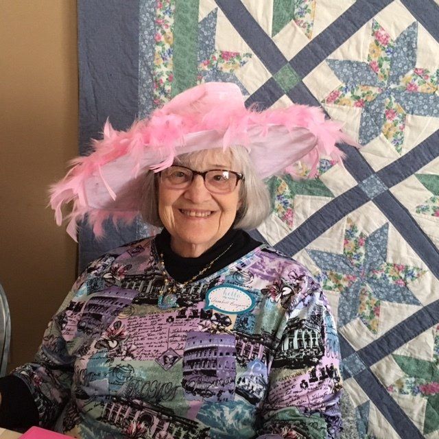 A woman wearing a pink hat and glasses is sitting in front of a quilt