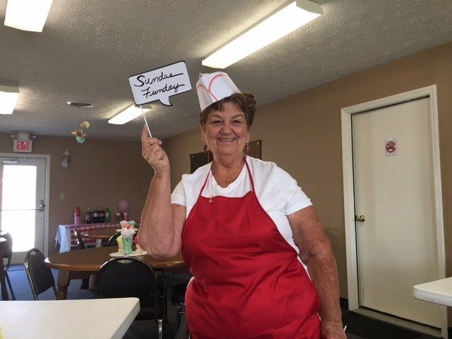 A woman in a red apron is holding a sign that says sunshine sundays