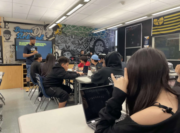 A group of people are sitting at tables in a classroom with graffiti on the wall