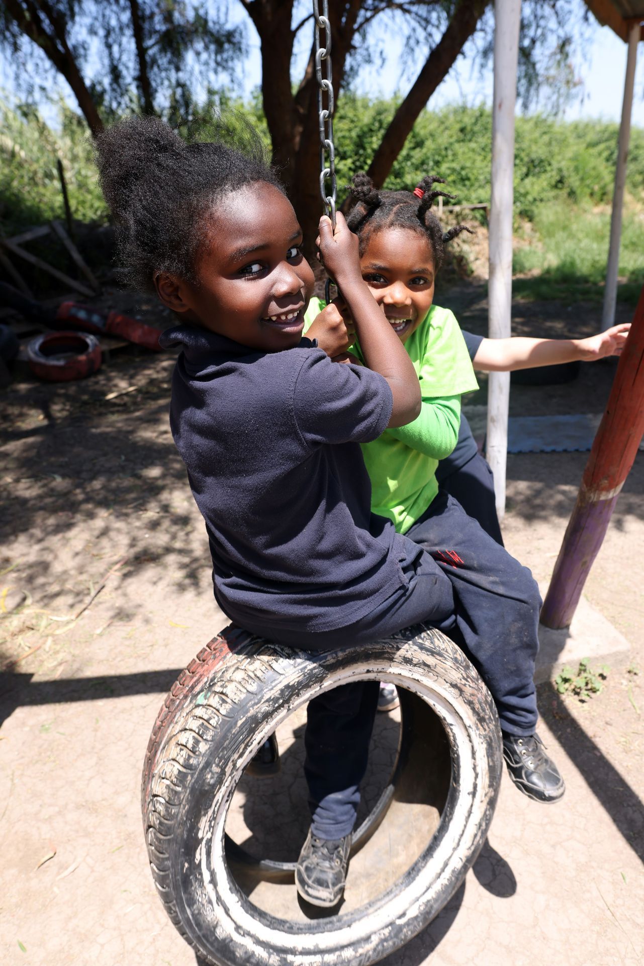 Dos niños se columpian en un columpio de neumático al aire libre; uno sonríe a la cámara, sujetando la cadena.