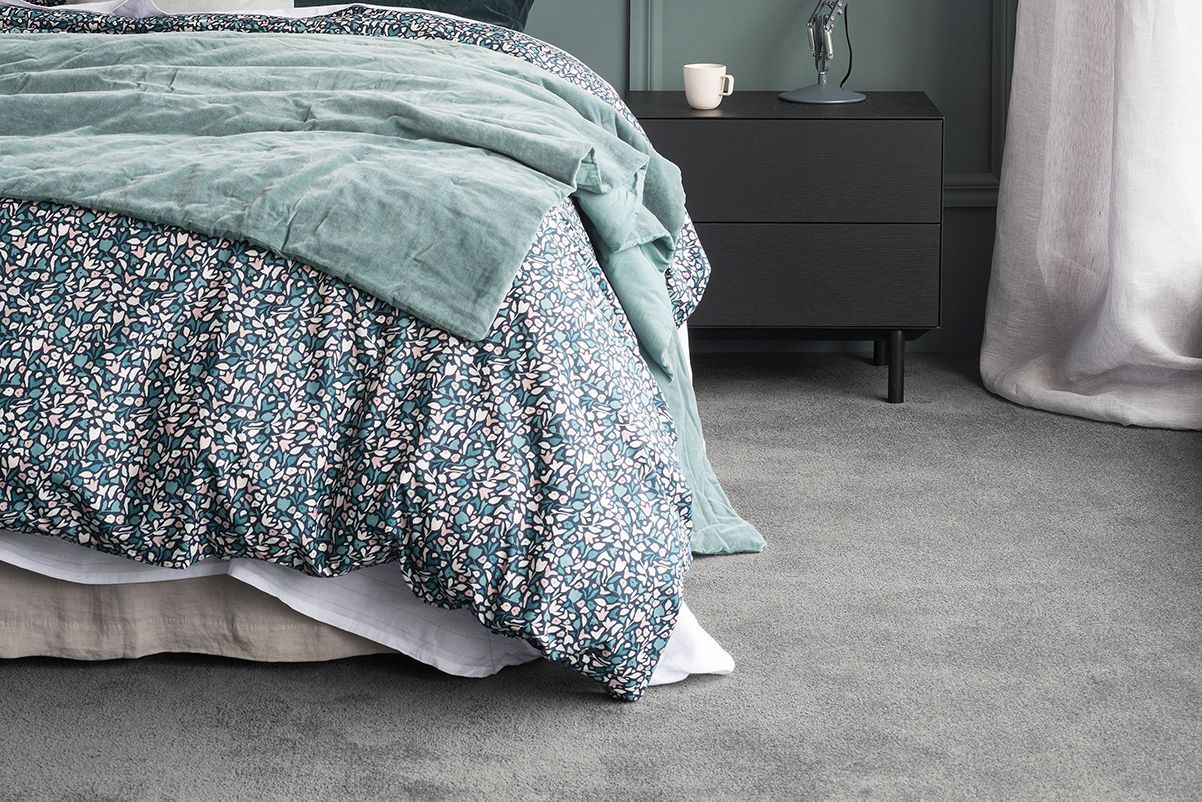 Bed with teal and floral bedding, gray carpet, and a black nightstand with a mug.— AGM Flooring & Tiling in Hidden Valley, QLD