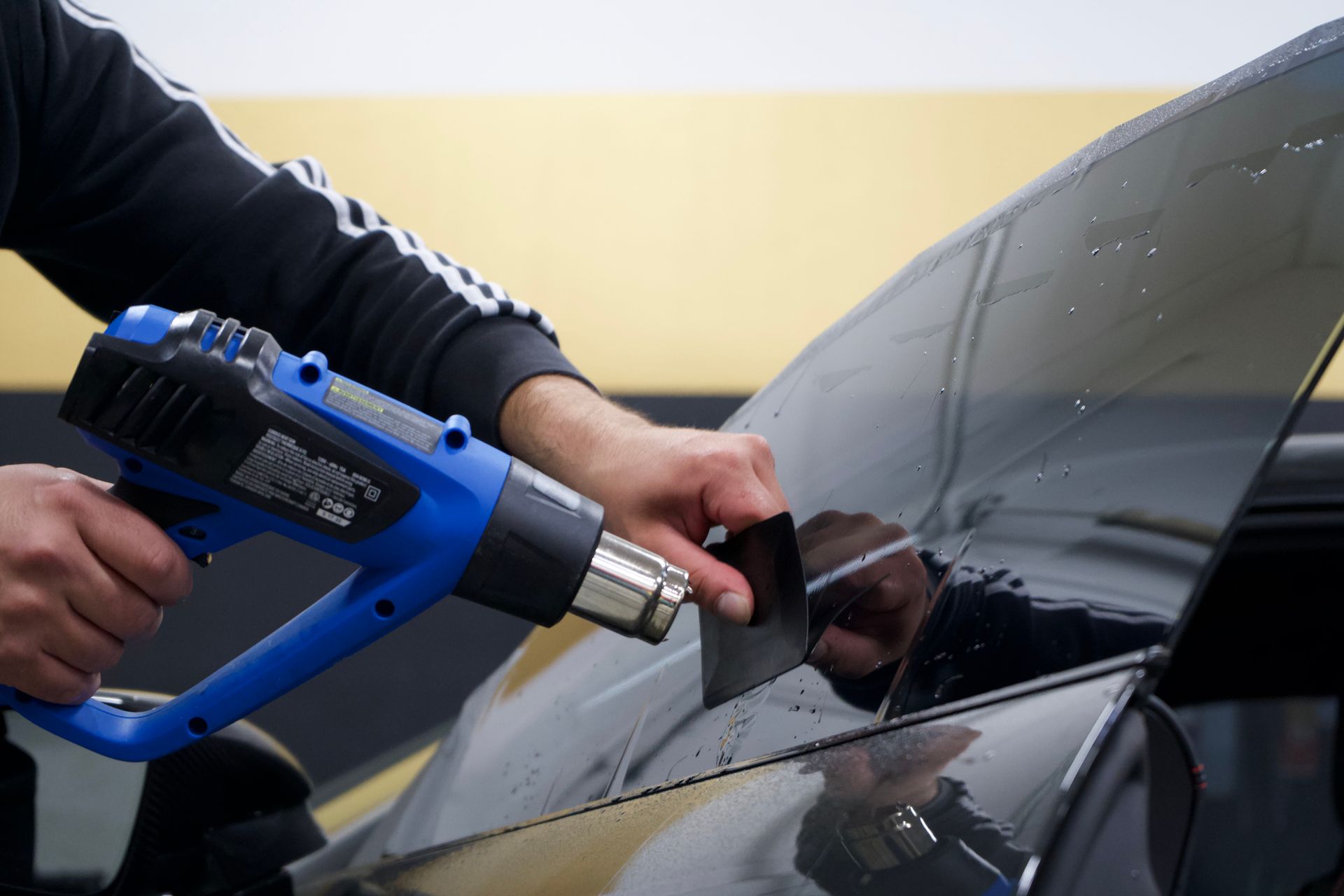 Person using a heat gun and squeegee to apply window tint to a car window.