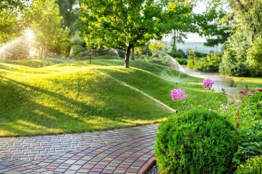 Sunlit park path beside green hills, trees, and pink flowers