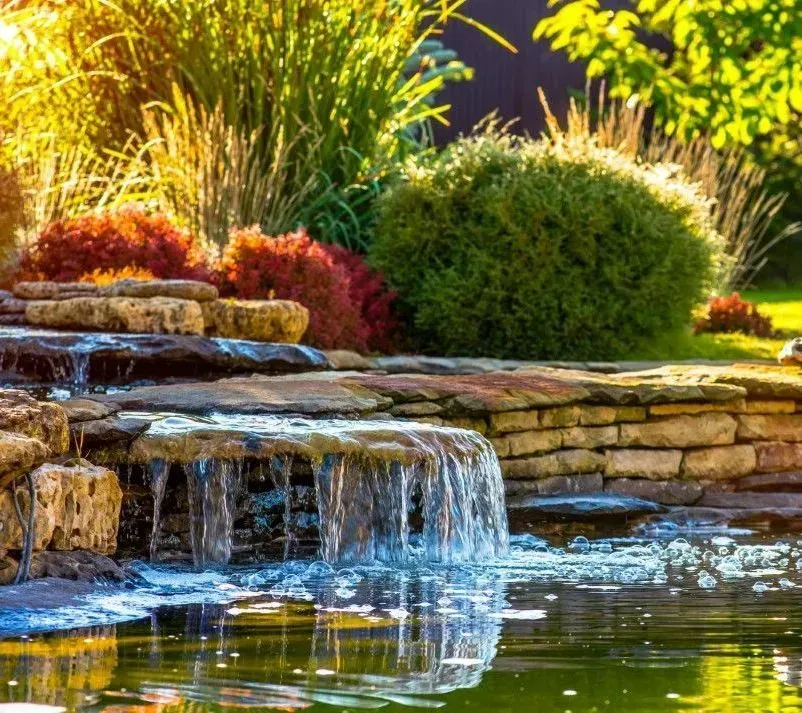 Stone garden waterfall with cascading water, lush green shrubs, and bright sunlight reflecting in the pond