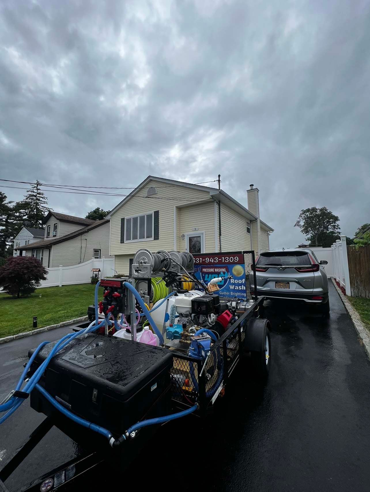 A house and a trailer with cleaning equipment, likely for pressure washing, parked on a driveway on an overcast day. A car is parked nearby.