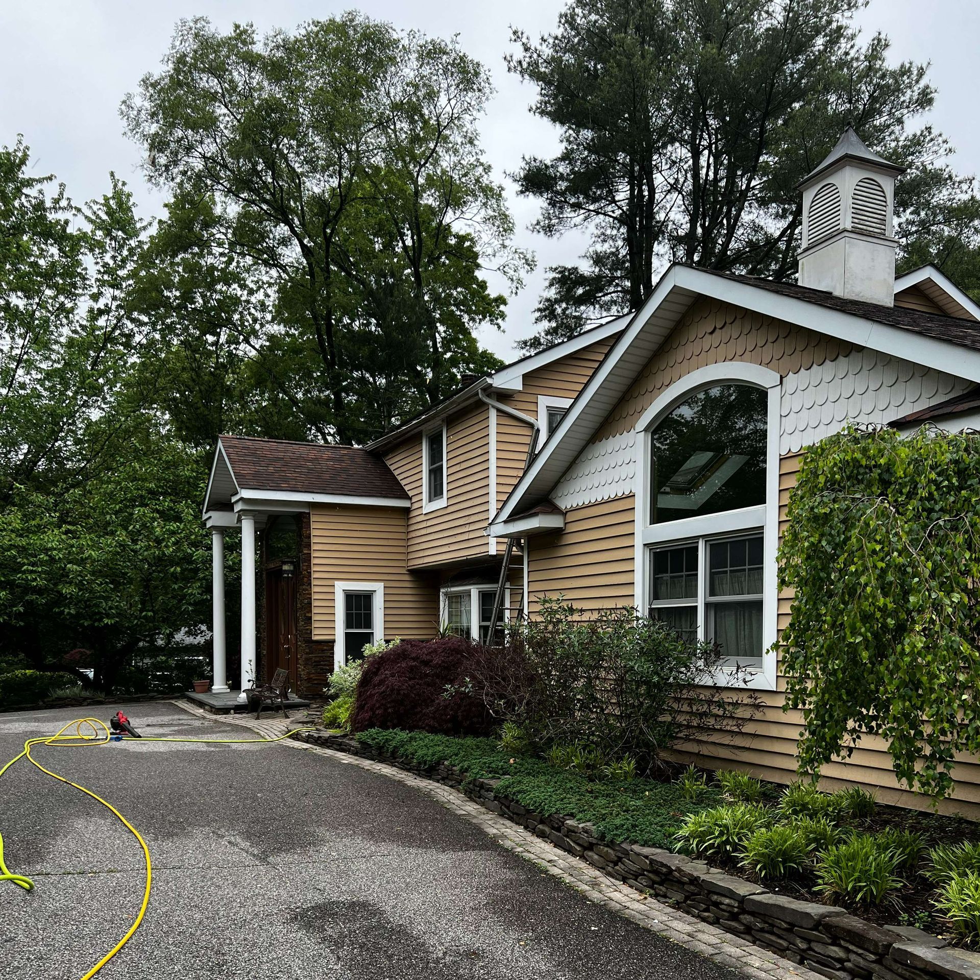 A two-story house with tan siding and white trim. Trees surround the home, and a yellow hose lies on the driveway.