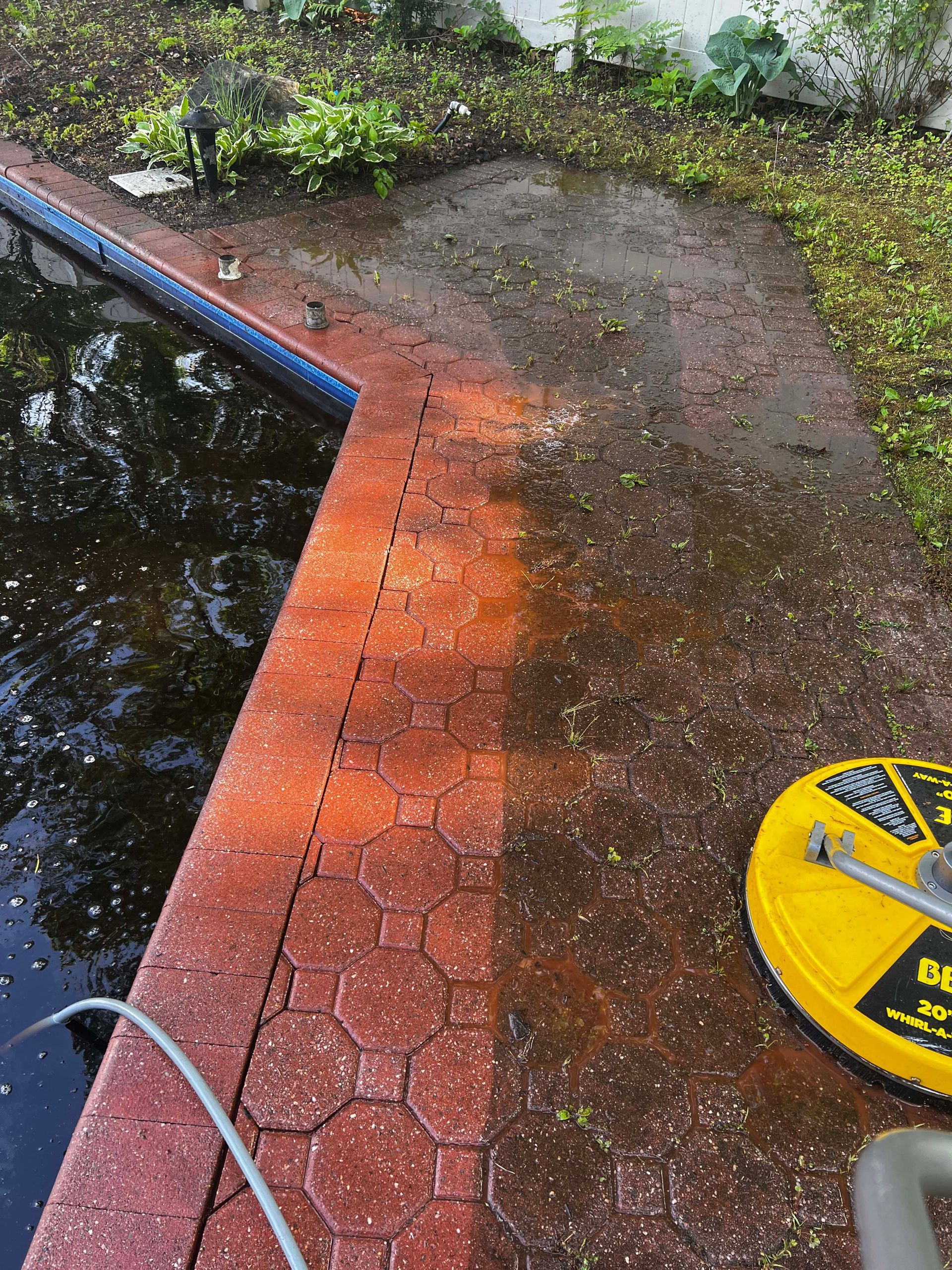 A person pressure washes a red brick patio near a swimming pool, revealing the cleaned surface.